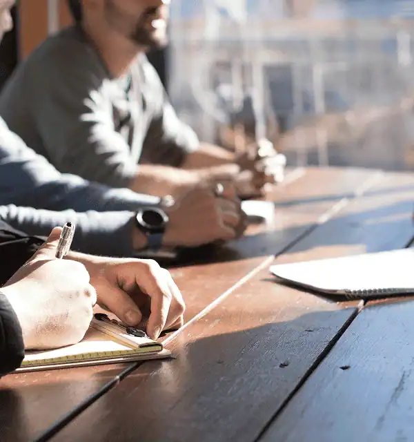 A professional taking notes by hand during a team session at a wooden table, with colleagues engaged in focused discussion illustrating Protea's intentional, belief-grounded approach to every client engagement.