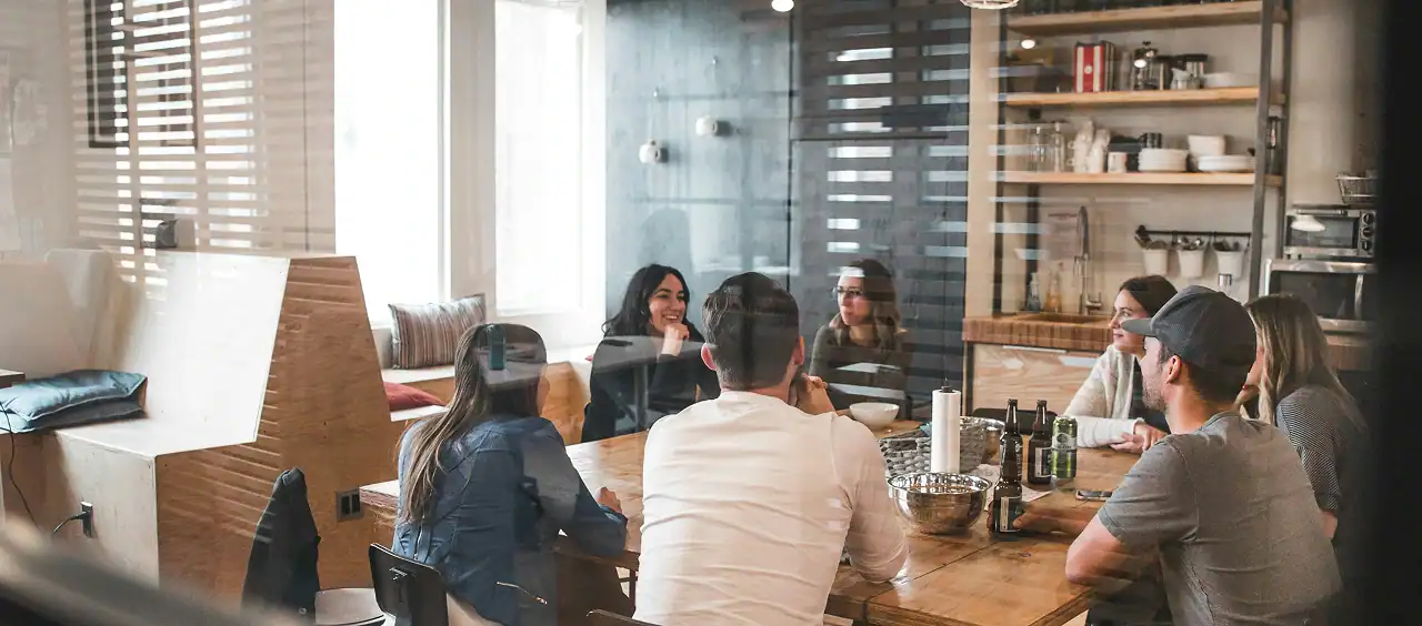 A diverse team of professionals gathered around a wooden table in a modern, light-filled workspace, engaged in open conversation reflecting Protea's belief-driven approach to collaboration and client engagement.