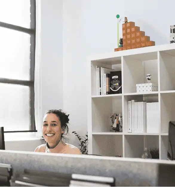 A smiling Protea team member at her desk in a bright, personality-filled office, with books and playful collectibles on a shelf behind her reflecting a workplace culture shaped by genuine values and human connection.