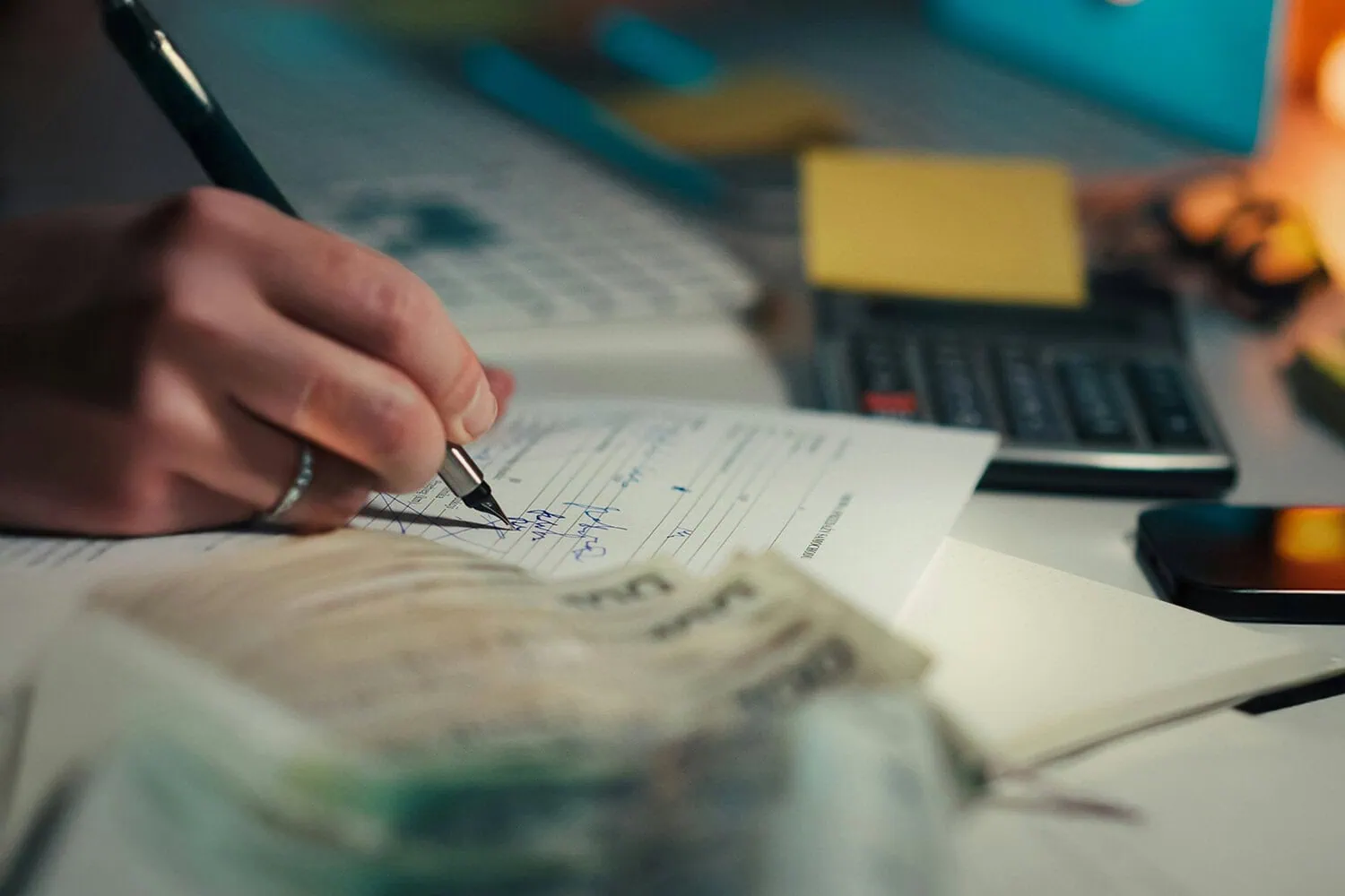 Hand signing financial documents with cash and calculator on desk